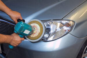 A person uses a power buffer to polish the headlight of a silver car.