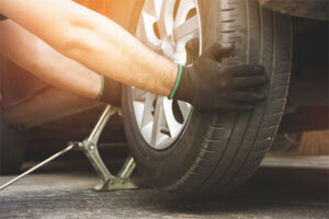 A man kneels beside a car, changing a flat tire with a wrench and jack in a roadside setting.