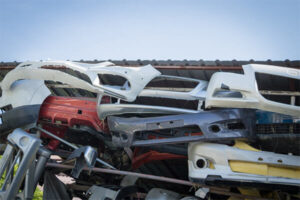 A pile of assorted used car bumpers stacked outdoors under a corrugated metal roof.